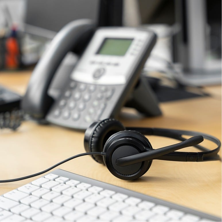 A black headset lies on a wooden desk next to a computer keyboard, with an office desk phone in the background.