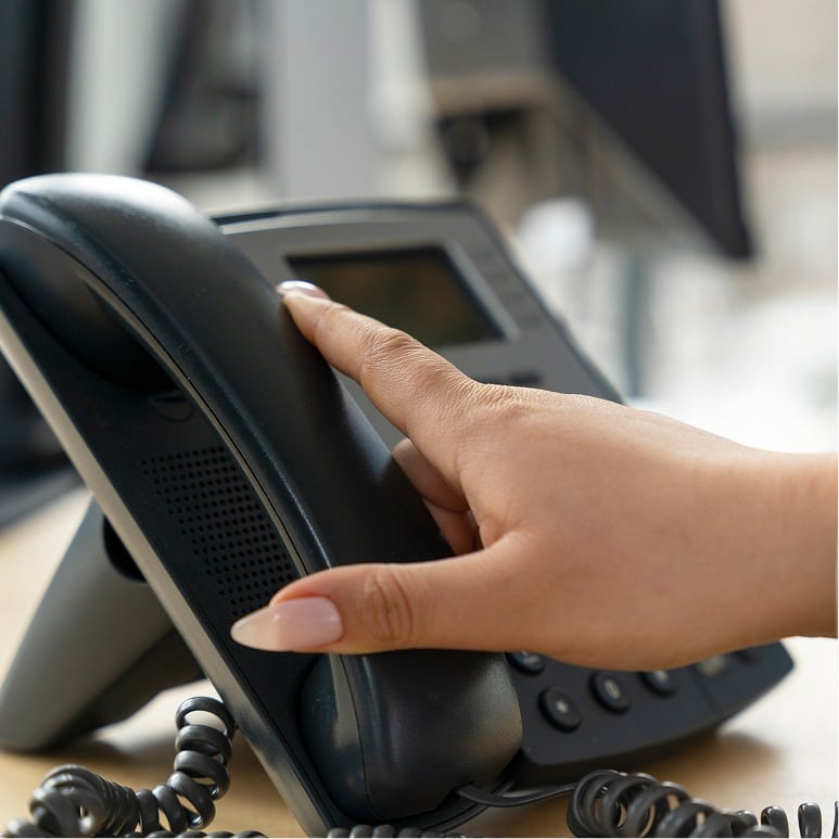 A person with manicured nails picks up the handset of a black office desk phone, with a coiled cord and buttons visible, in a modern office setting.
