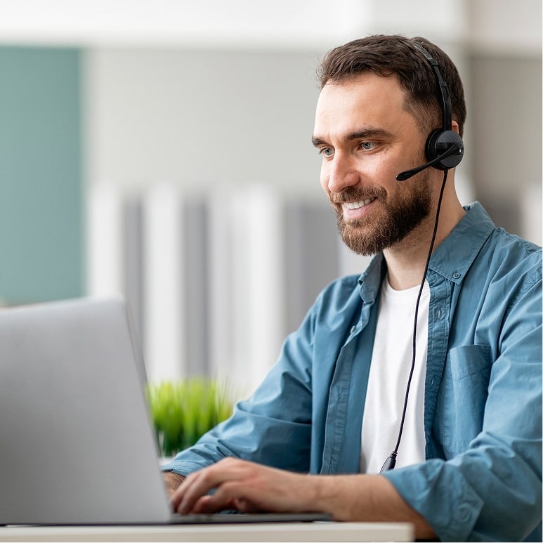 A man wearing a headset smiles while working on a laptop at a desk in a bright, modern office setting.