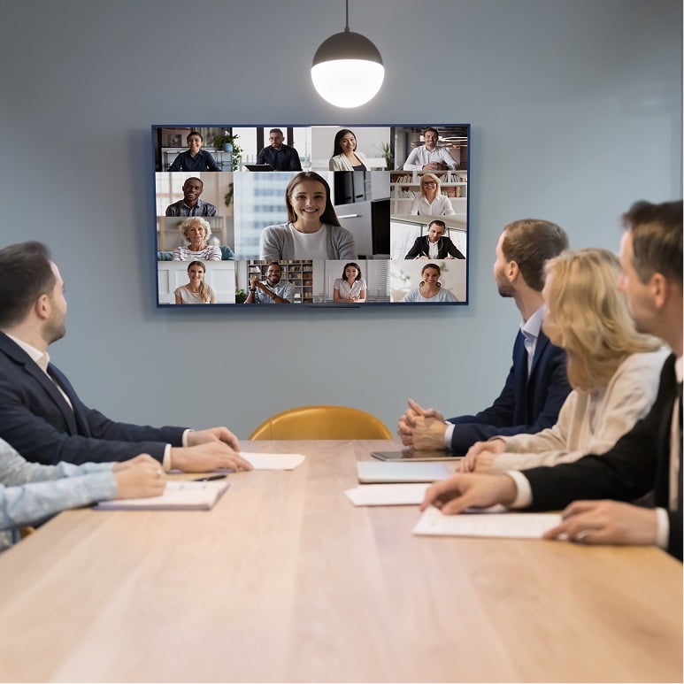 Five people sit at a conference table watching a large screen showing a video call with multiple participants. The room is modern, with blue walls and a hanging light above the table.
