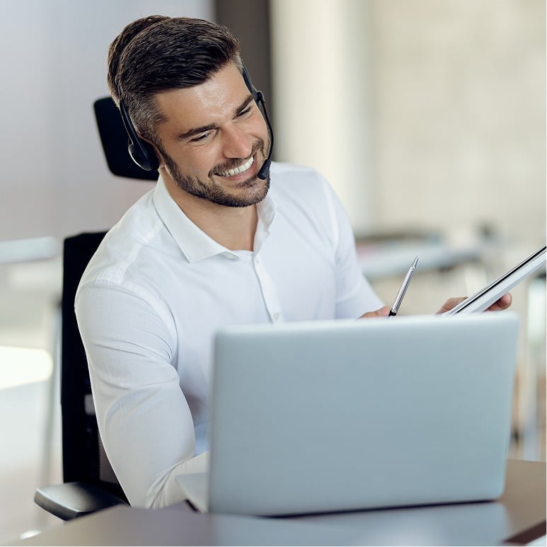 A smiling man wearing a headset sits at a desk with a laptop, holding a pen and notepad, appearing engaged in a video call or virtual meeting in a bright office setting.