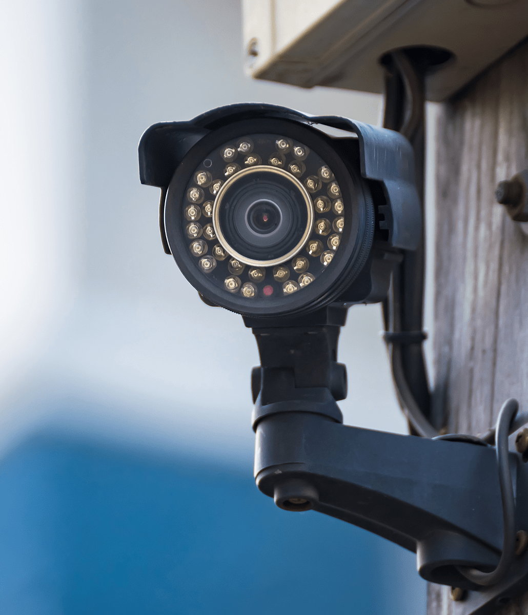 A close-up of a black security camera mounted on a wooden pole, with visible infrared LEDs surrounding the lens. The background is blurred with blue and white tones.