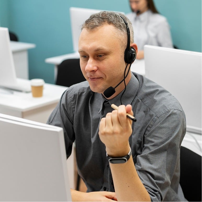 A man wearing a headset sits at a desk in an office, looking at a computer monitor and holding a pen. Another person is visible in the background, also working at a desk.