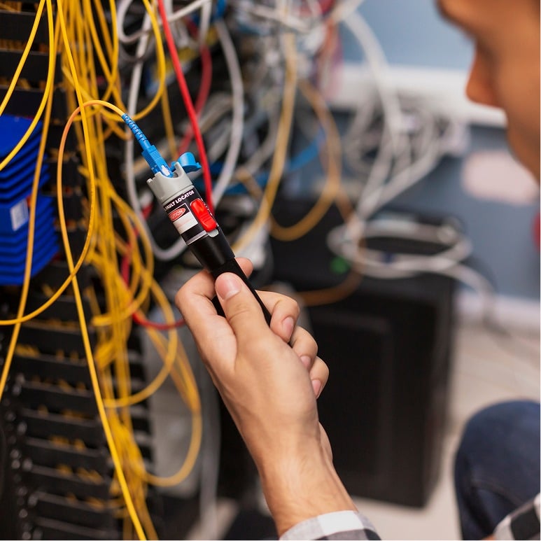 A person uses a handheld network cable tester to check yellow fiber optic cables in a server room, with numerous wires and equipment visible in the background.