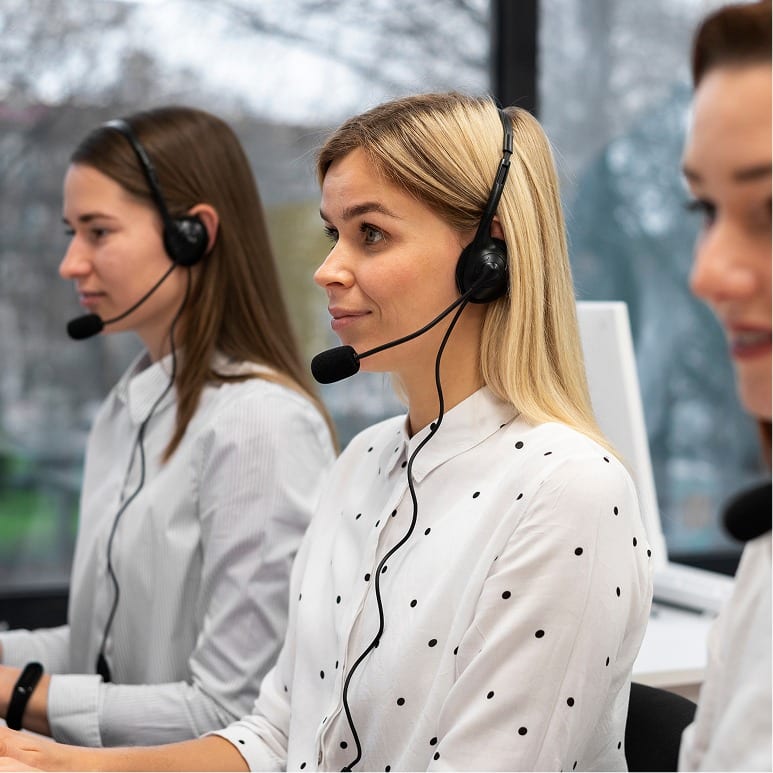 Three women wearing headsets and white shirts sit in a row at desks, working as customer service representatives in a bright office with large windows in the background.