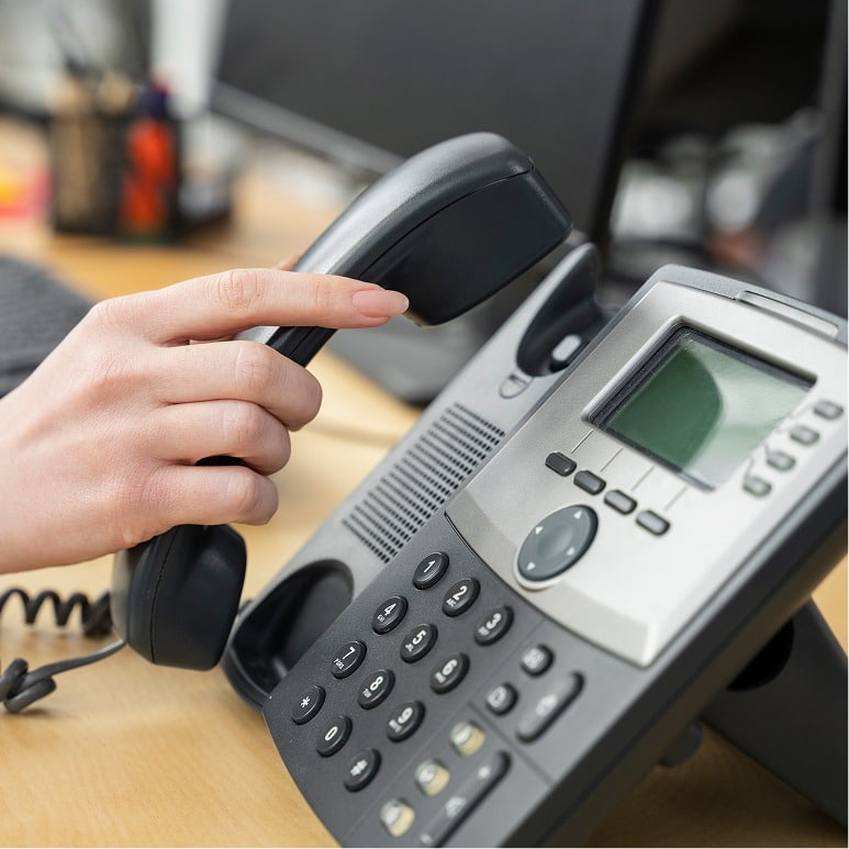 A person’s hand picking up the receiver of an office desk phone, with a computer monitor and office supplies blurred in the background.