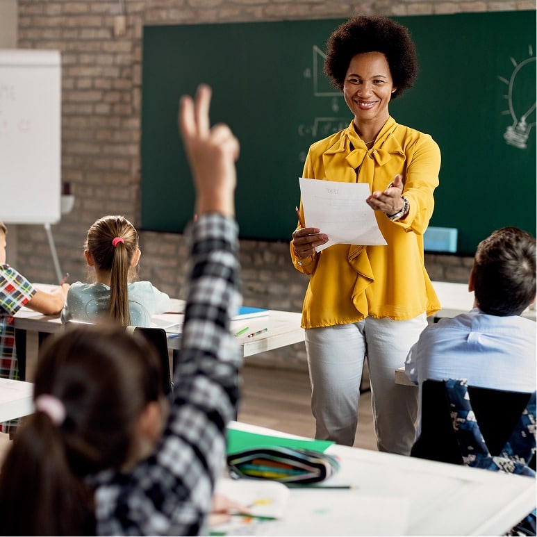 A smiling teacher in a yellow blouse stands at the front of a classroom, holding papers and gesturing towards a student raising their hand. Several children sit at desks facing the teacher and a chalkboard.