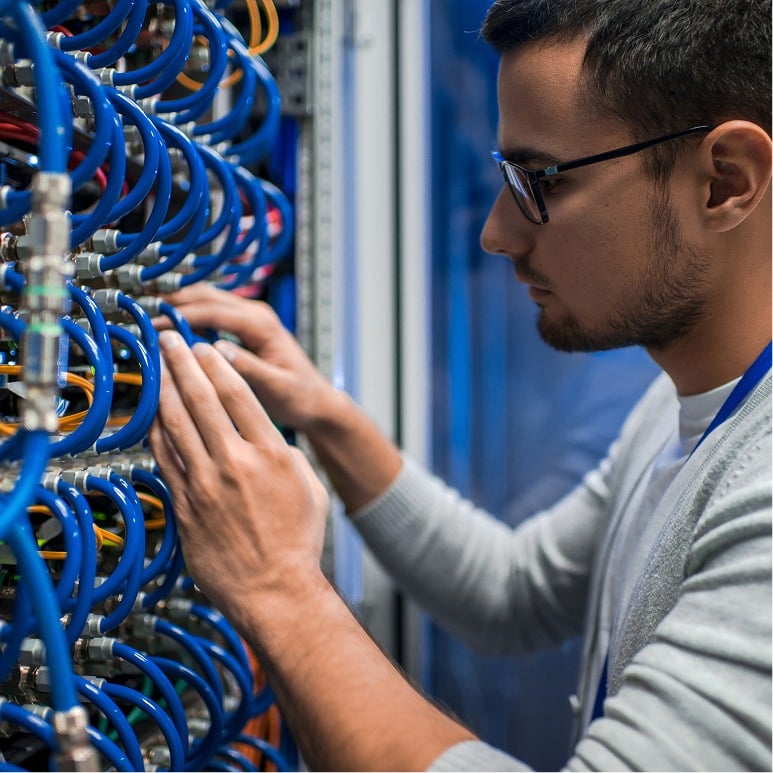 A person wearing glasses works in a server room, connecting or inspecting blue network cables organized in a rack.