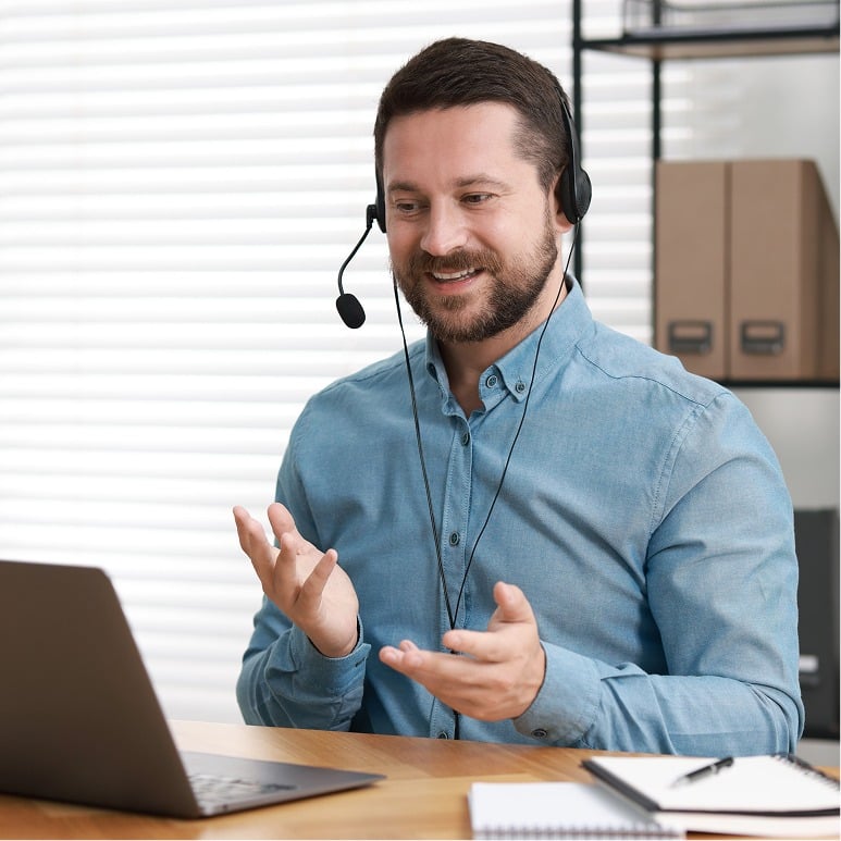 A man wearing a headset and a blue shirt sits at a desk, smiling and gesturing with his hands while having a video call on his laptop in a bright, modern office setting.