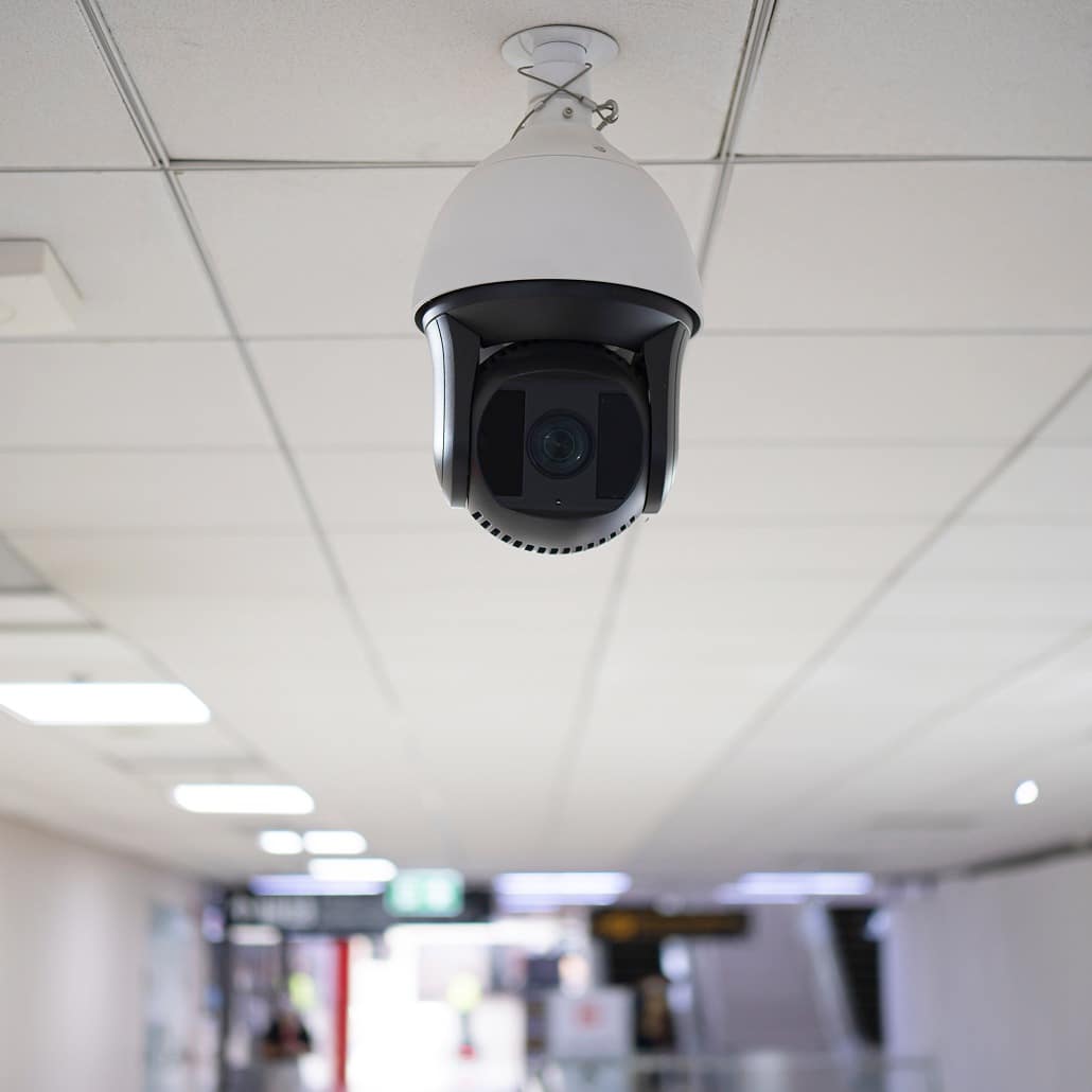A security camera is mounted on a white ceiling in a well-lit indoor area, monitoring a hallway or corridor with signs and blurred objects visible in the background.