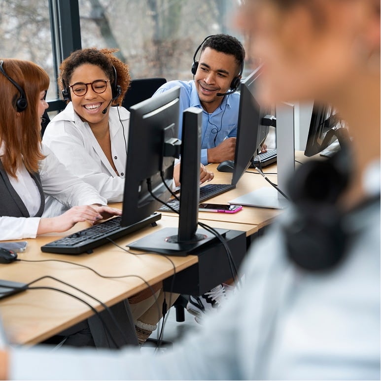 Three people wearing headsets sit at desks with computers, smiling and talking to each other in a modern office. Another person in the foreground is out of focus. The scene suggests a call center or collaborative work environment.
