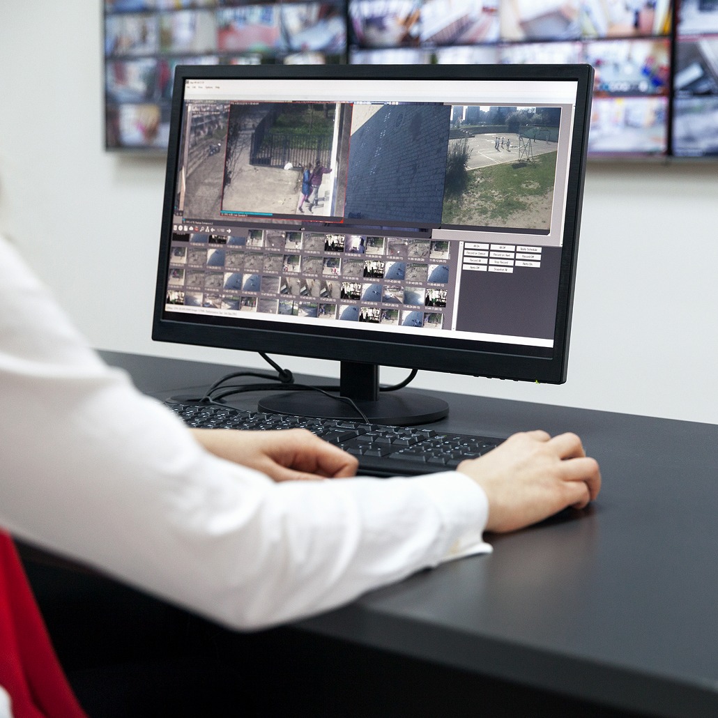 A person sits at a desk monitoring multiple live security camera feeds on a computer screen in a surveillance control room.