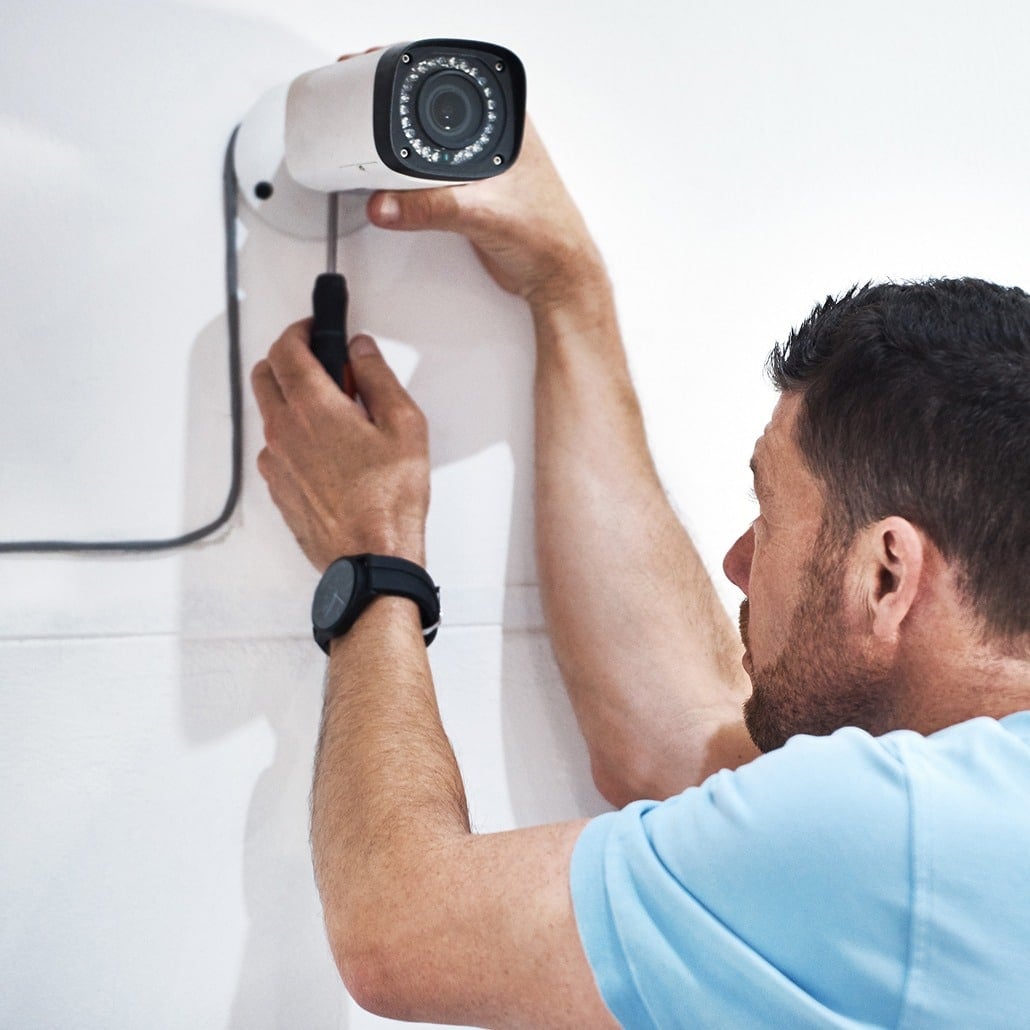 A man in a light blue shirt installs a security camera on a white wall, using a screwdriver to secure the device and adjust its position.