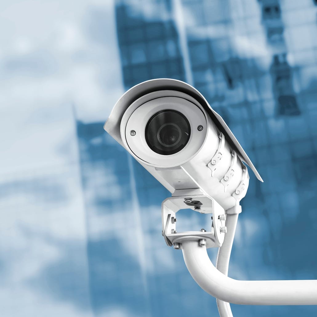 A close-up of a security camera mounted on a white bracket, with a blurred blue and glass building in the background.