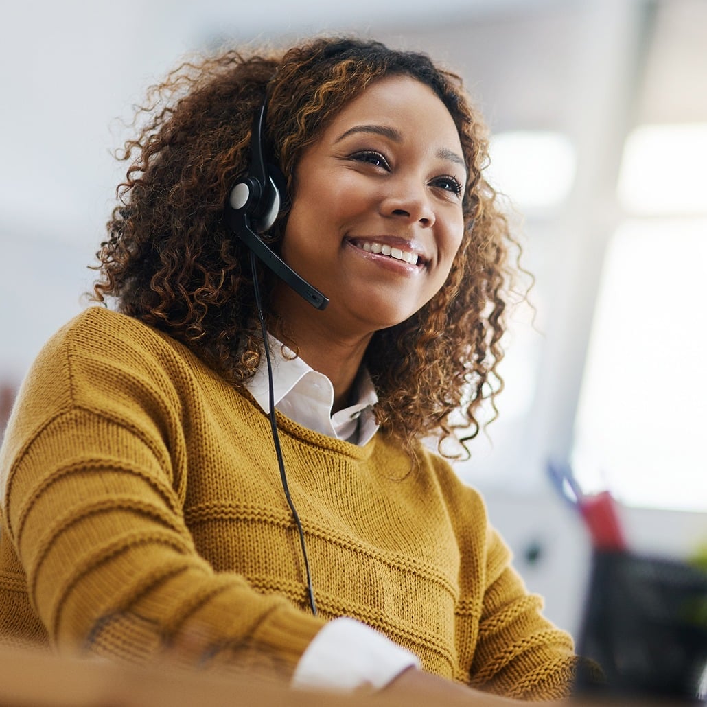 A woman with curly hair, wearing a mustard yellow sweater and a headset, smiles while working at a desk in a bright office environment.