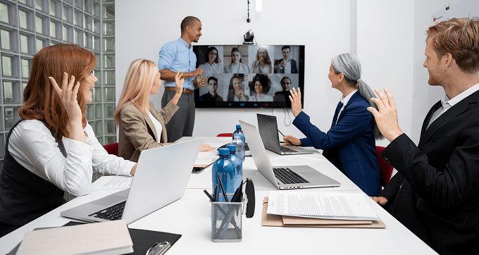 Five people in a conference room wave at a screen showing a video call with multiple participants. Laptops, documents, and water bottles are on the table. One person stands and speaks to the group.