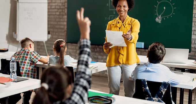 A smiling teacher holding papers gestures to a student raising their hand in a classroom, with other students seated at desks facing a chalkboard with drawings.