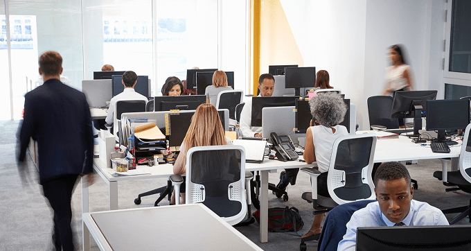 Open-plan office with several people working at desks with computers; some are seated and focused on their screens, while two people walk through the workspace. The atmosphere appears busy and professional.