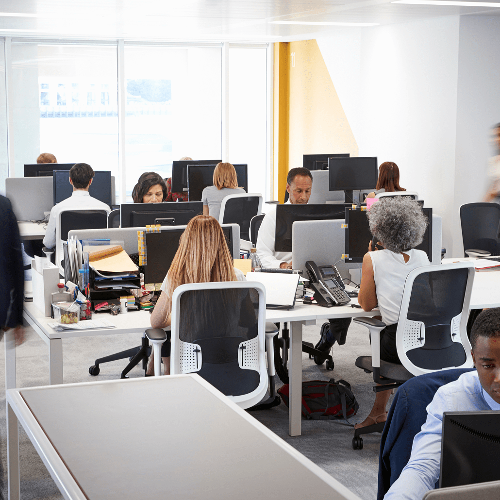 Open-plan office with several people working at desks with computers, papers, and office supplies. Some employees are focused on screens; others are talking or walking. The space is modern, bright, and has large windows.