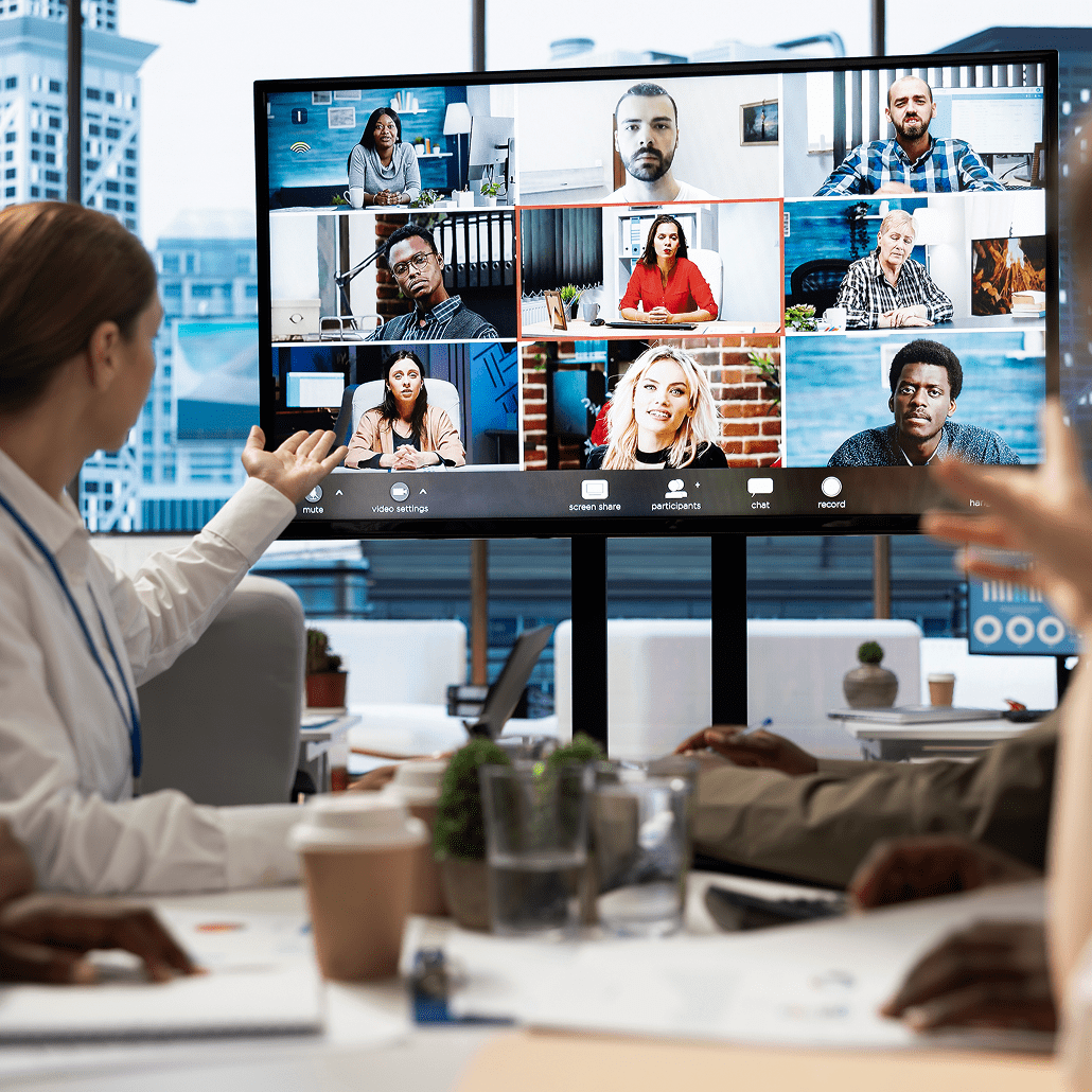 A person in an office meeting room gestures toward a large screen displaying a video conference with nine diverse participants in separate windows. Laptops, documents, and coffee cups are on the table.