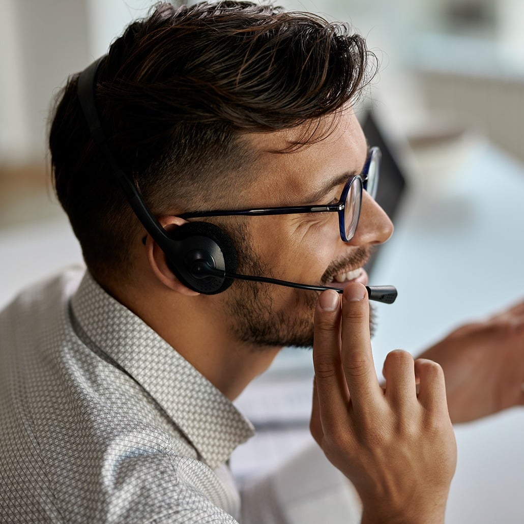 A man wearing glasses and a headset smiles while speaking, possibly during a video call or customer support interaction, sitting at a desk in a bright office.