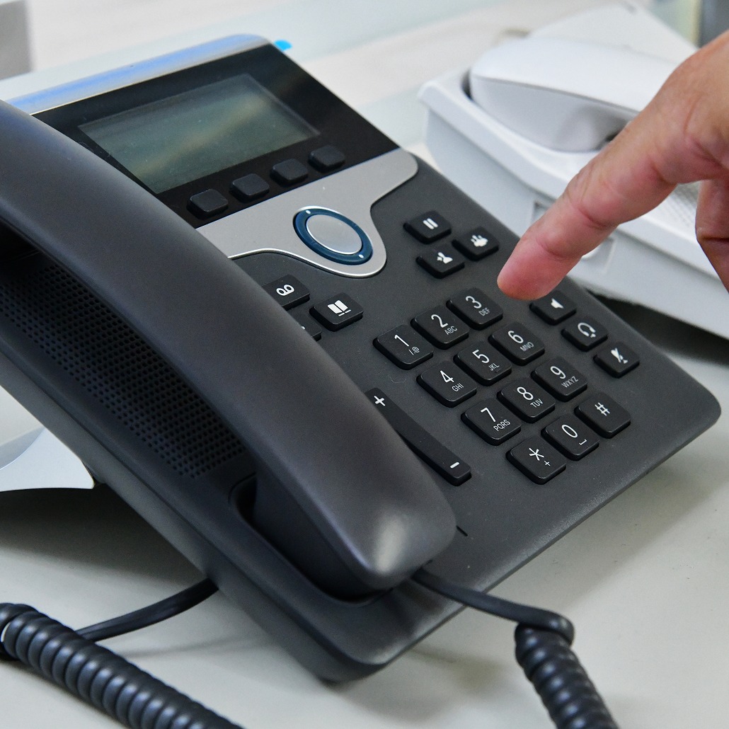 A close-up of a person’s finger pressing a button on a black office desk phone with a digital display and a corded handset. Another white phone is visible in the background.