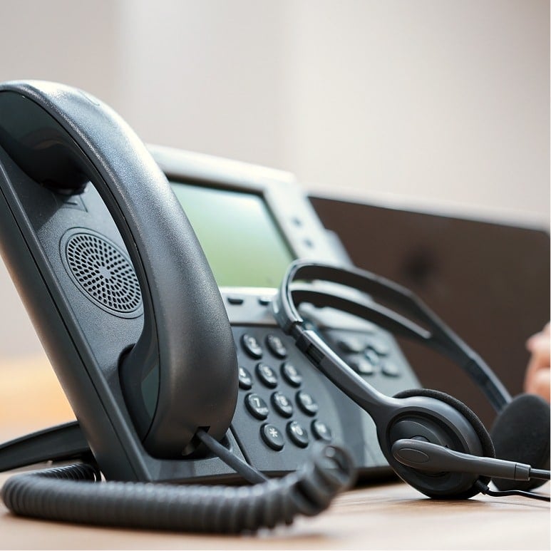 A close-up of a black office desk phone with a corded handset and a headset resting beside it on a wooden surface. The background is softly blurred.