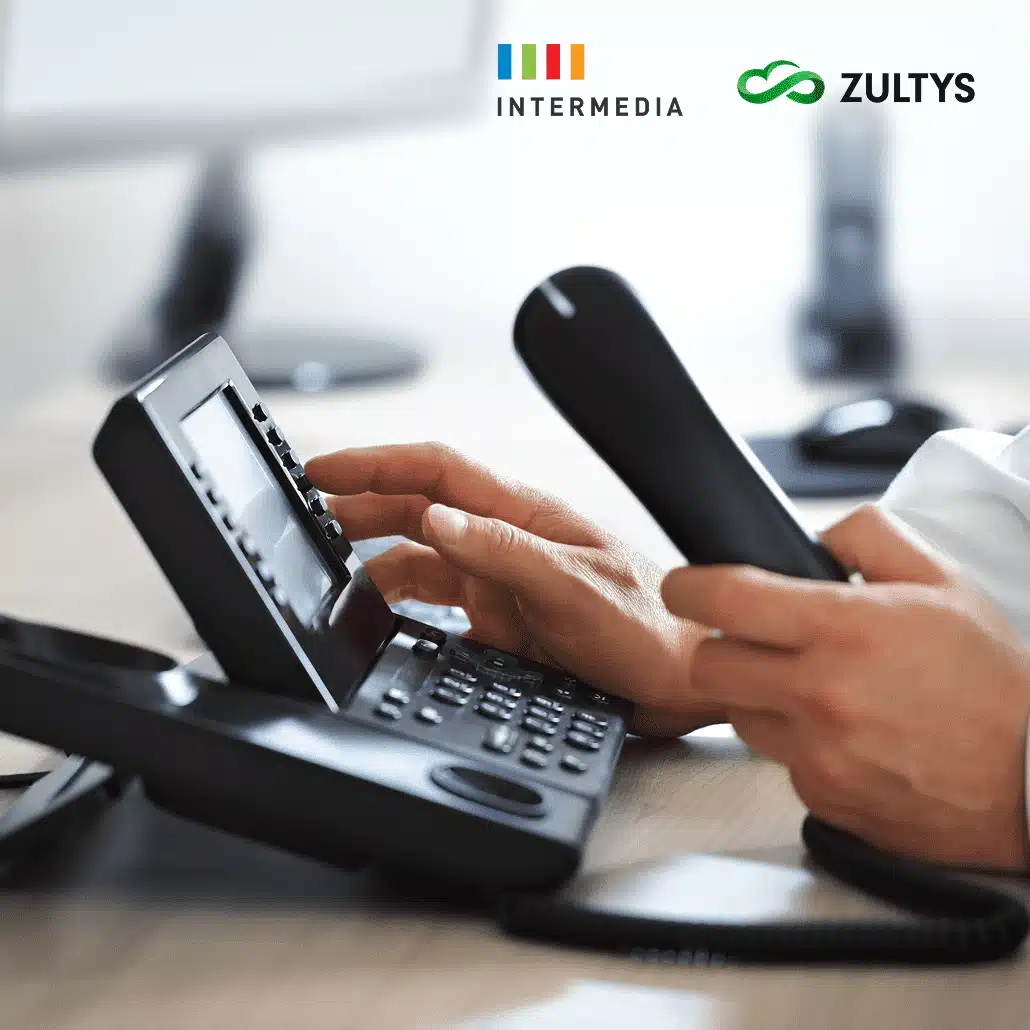 A person dials a number on an office desk phone while holding the receiver. The logos of Intermedia and Zultys are visible in the top right corner of the image.