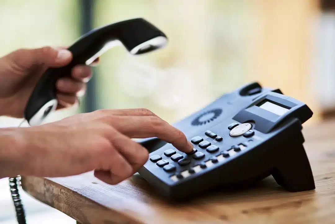 A person holds a telephone handset in one hand while dialing a number on a black landline phone with the other hand, positioned on a wooden surface.