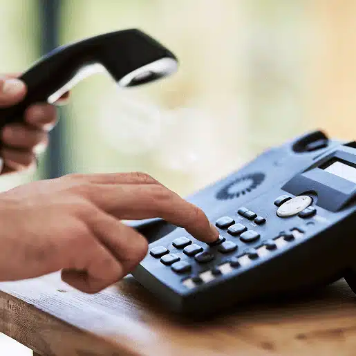 A person holds a telephone receiver in one hand and dials a number on a black landline phone with the other hand, set on a wooden surface.
