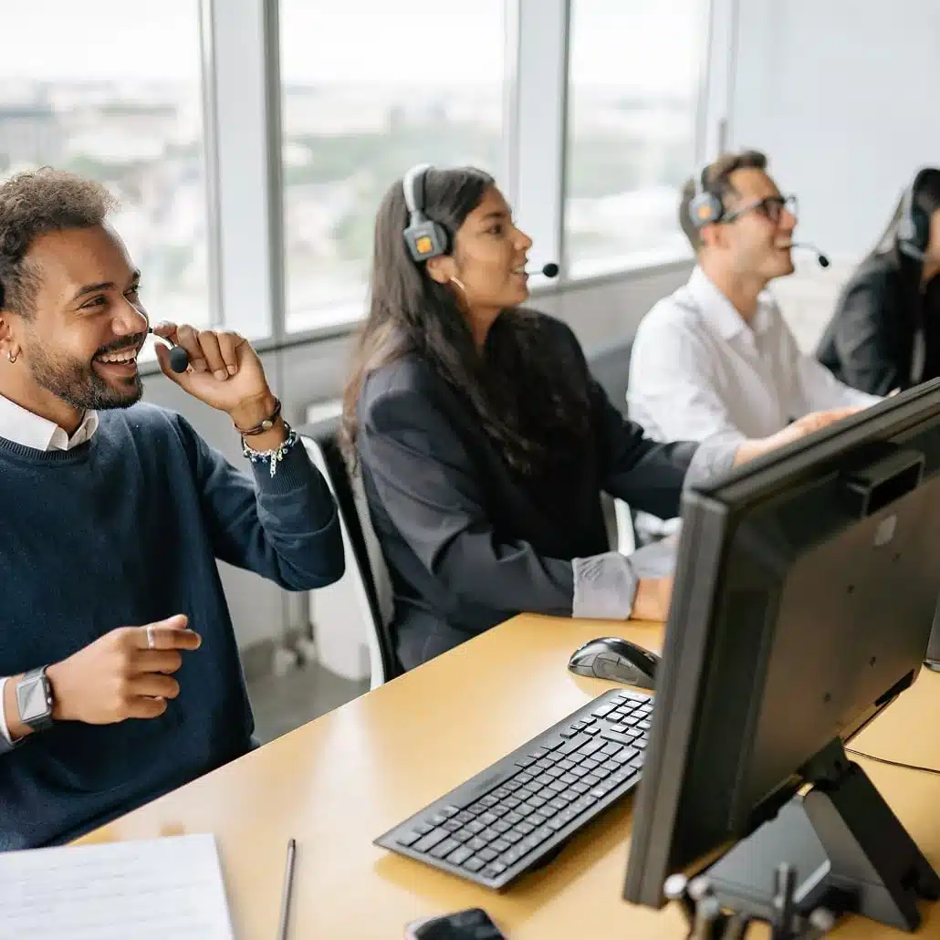 Three people wearing headsets sit at a desk with computers in a bright office, smiling and talking, appearing to work in a customer service or call center environment.