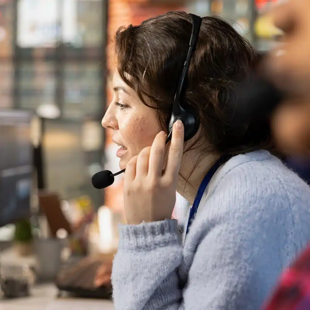A woman wearing a headset and speaking into a microphone works at a computer in an office setting.