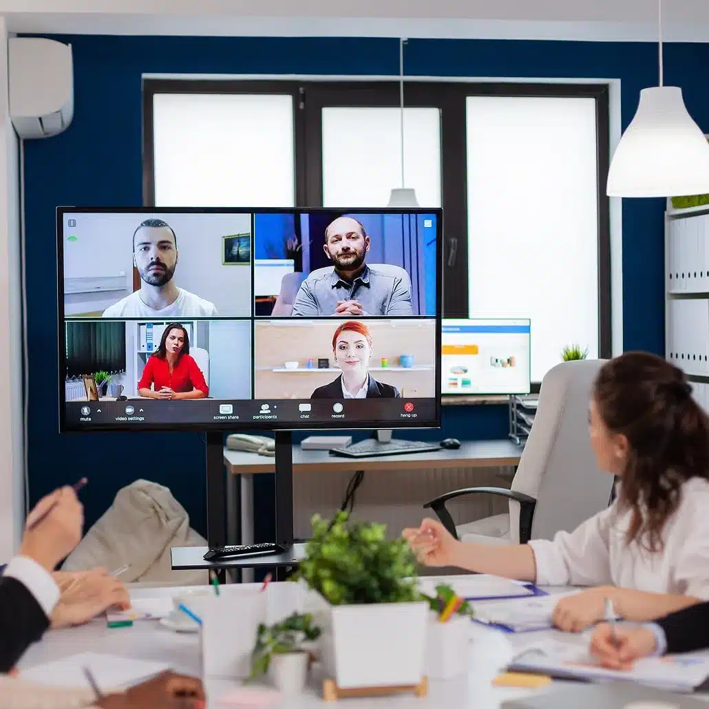 A group of people in an office sit around a table watching a video conference on a large monitor, which displays five participants in a virtual meeting. Papers and laptops are on the table.