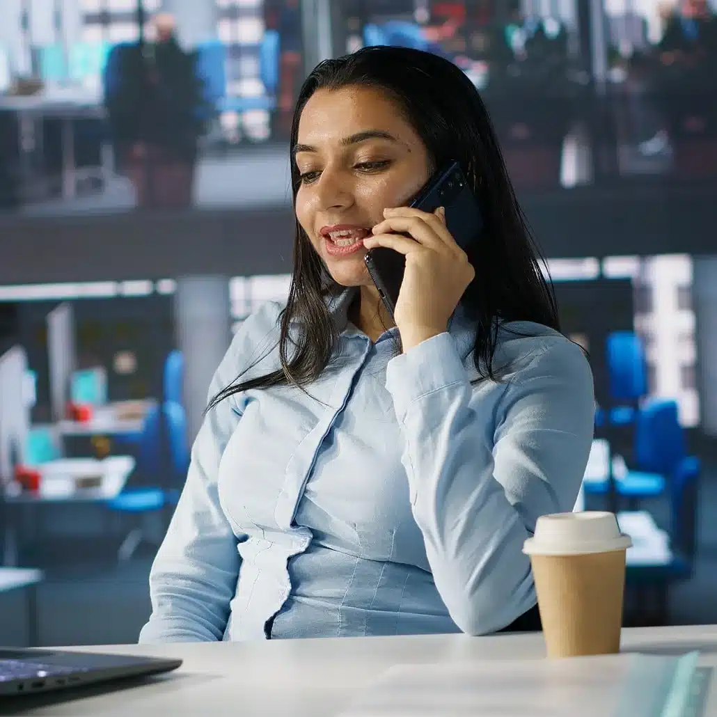 A woman in a light blue blouse talks on a smartphone while sitting at a desk with a laptop and a takeaway coffee cup in a modern office setting.