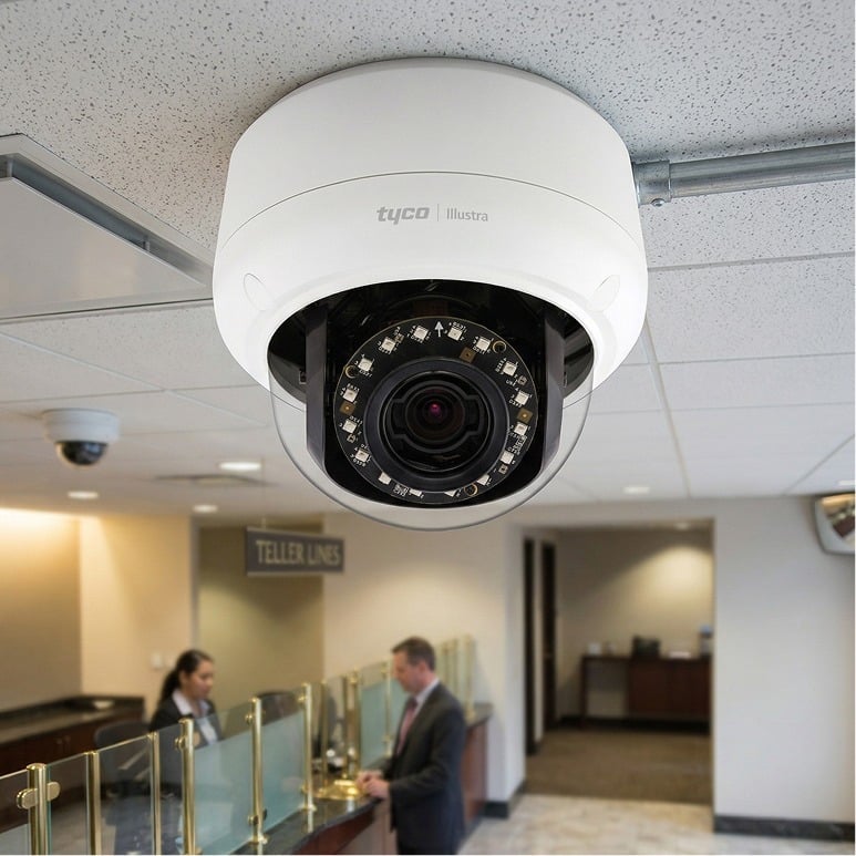 A close-up of a ceiling-mounted security camera in a bank lobby, with two people speaking at the teller counter in the background. Signs read "TELLER LINES." The camera is labeled "tyco Illustra.