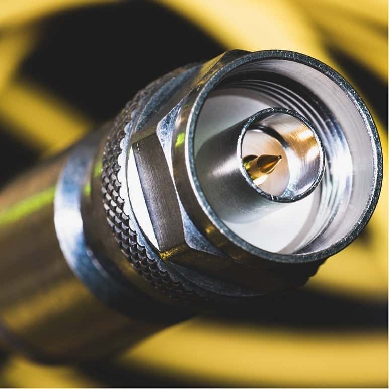 Close-up of a coaxial cable connector with a metallic finish, showing the central pin and threaded outer shell. Yellow cables are blurred in the background.