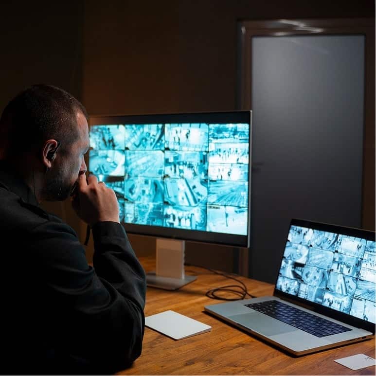 A person sits at a desk, monitoring multiple security camera feeds displayed on a large computer monitor and a laptop in a dimly lit room. A closed door is visible in the background.