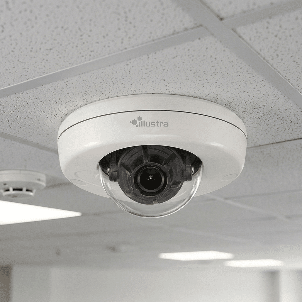 A white dome security camera is mounted on a ceiling with white acoustic tiles. The camera lens is visible inside the clear dome cover, and part of a smoke detector is also seen nearby.
