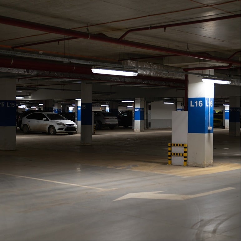 An underground parking garage with several parked cars and blue signs marking spaces L15 and L16, lit by overhead fluorescent lights. An arrow is painted on the floor, and a yellow-black barrier is near a column.