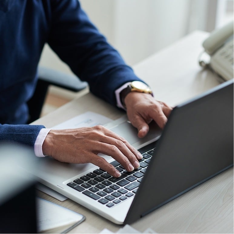 A person wearing a dark blue sweater and gold watch types on a laptop at a desk with papers and a phone nearby.