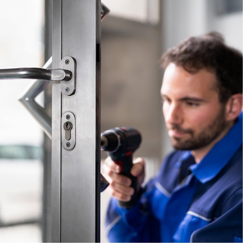 A locksmith in a blue uniform uses a power drill to repair or install a lock on a metal door, with the focus on the lock and the worker blurred in the background.