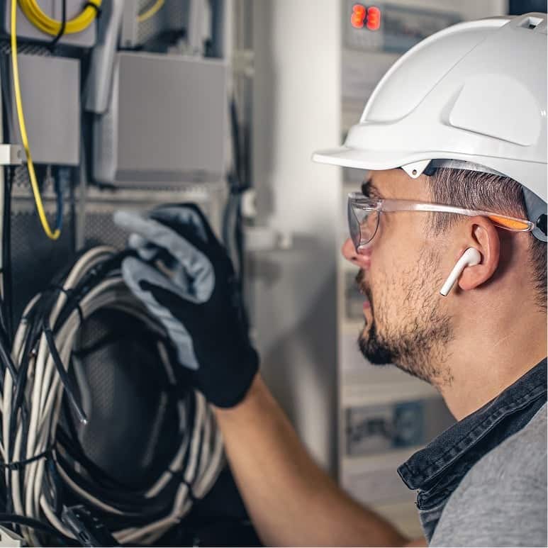 A man wearing a white hard hat, safety glasses, gloves, and earbuds inspects electrical cables and equipment inside a control panel or server box.