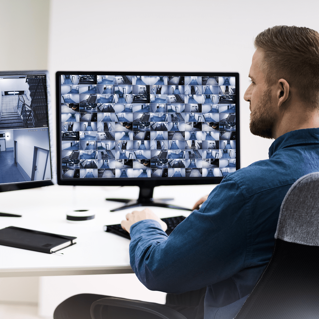 A person in a blue shirt sits at a desk monitoring multiple surveillance camera feeds on two computer screens in a modern office setting. A notebook and a wireless device are also on the desk.
