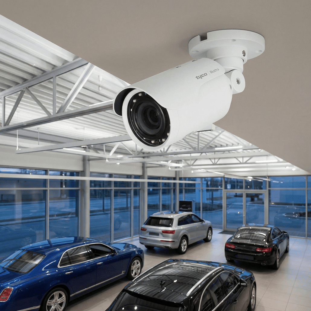 A security camera is mounted on the ceiling of a modern car showroom, overlooking several luxury vehicles parked inside near large glass windows.