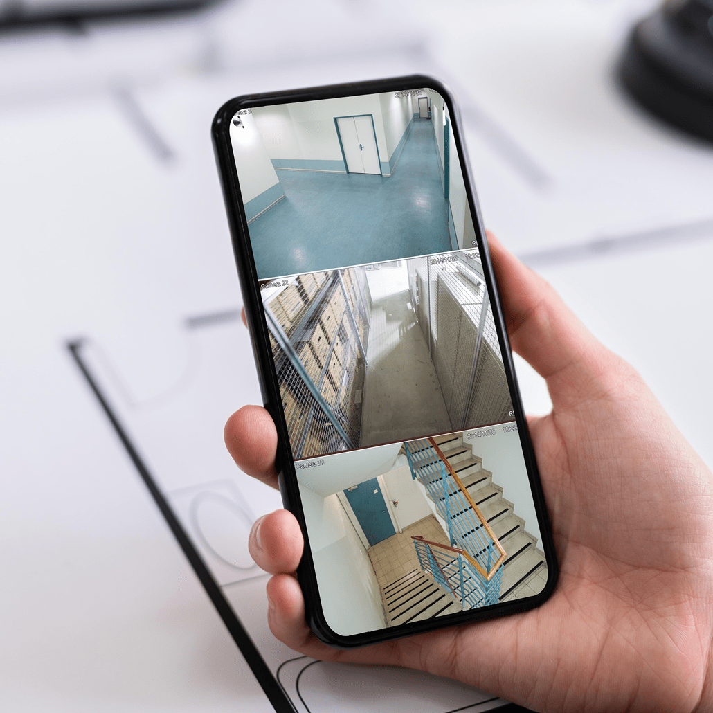 A hand holds a smartphone displaying four security camera feeds, showing views of hallways, a stairwell, and storage areas inside a building.