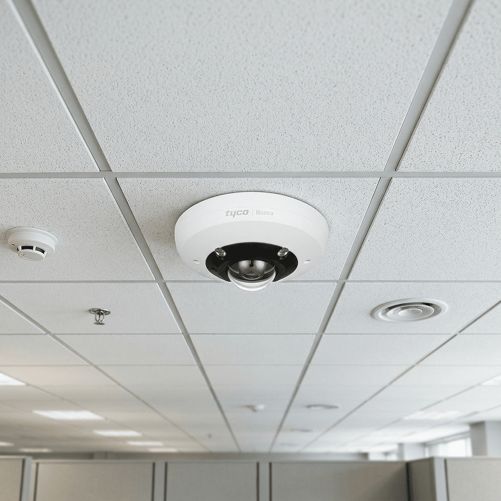 A white security camera is mounted on a ceiling with white tiles in an office setting. Nearby, there is a smoke detector and an air vent, with cubicle walls visible below.